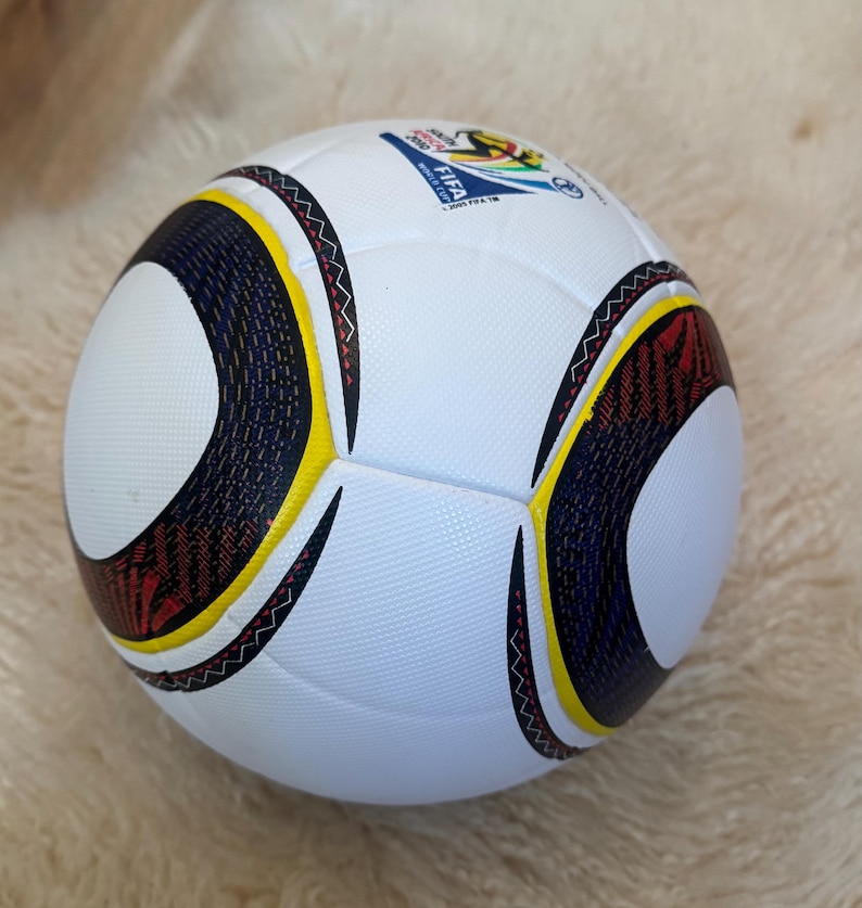 K&ouml;nnte beinhalten: Wei&szlig;er Fu&szlig;ball mit schwarzen, gelben und roten geschwungenen Akzenten. Mit dem Logo der FIFA Fussball-Weltmeisterschaft 2006. Der Ball hat eine strukturierte Oberfl&auml;che.