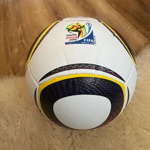 K&ouml;nnte beinhalten: Ein wei&szlig;-schwarzer Fu&szlig;ball mit gelben und marineblauen Akzenten. Der Ball zeigt das FIFA World Cup South Africa 2010 Logo. Es ist ein offizieller Spielball.