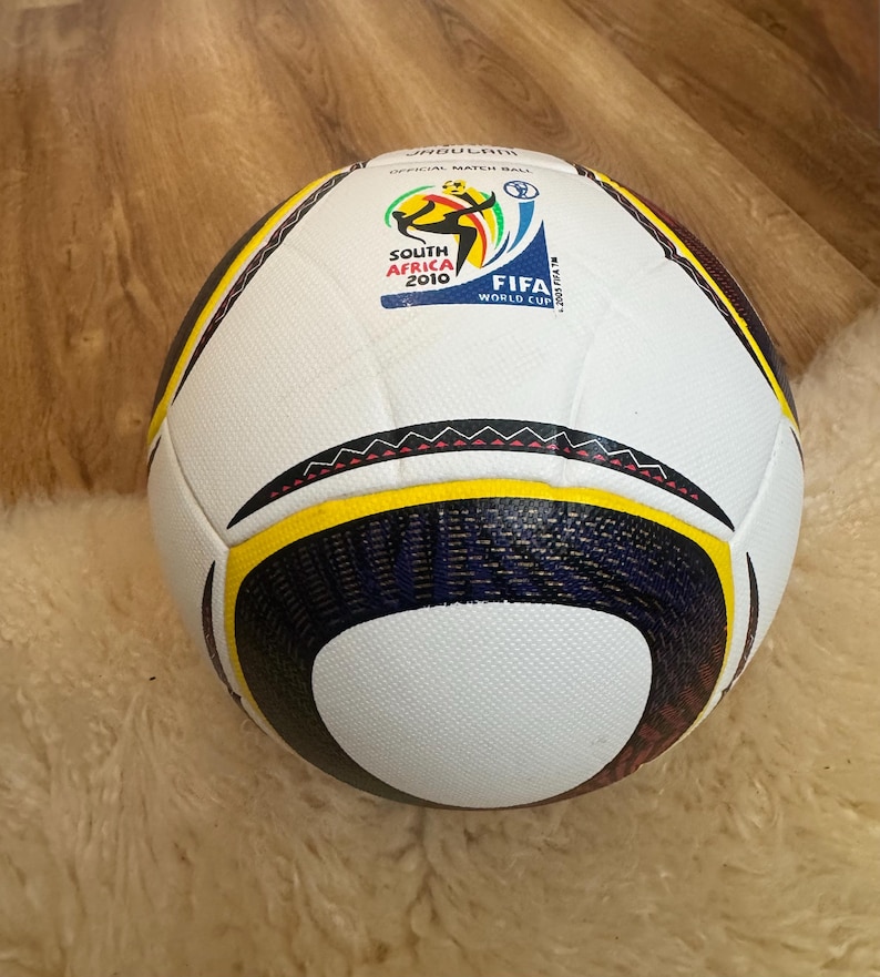 K&ouml;nnte beinhalten: Ein wei&szlig;-schwarzer Fu&szlig;ball mit gelben und marineblauen Akzenten. Der Ball zeigt das FIFA World Cup South Africa 2010 Logo. Es ist ein offizieller Spielball.