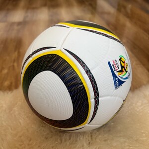 K&ouml;nnte beinhalten: Ein wei&szlig;er Fu&szlig;ball mit schwarzen, gelben und roten Akzenten. Der Ball hat eine strukturierte Oberfl&auml;che und ein Design mit dem Text "SOUTH AFRICA 2010 FIFA WORLD CUP".