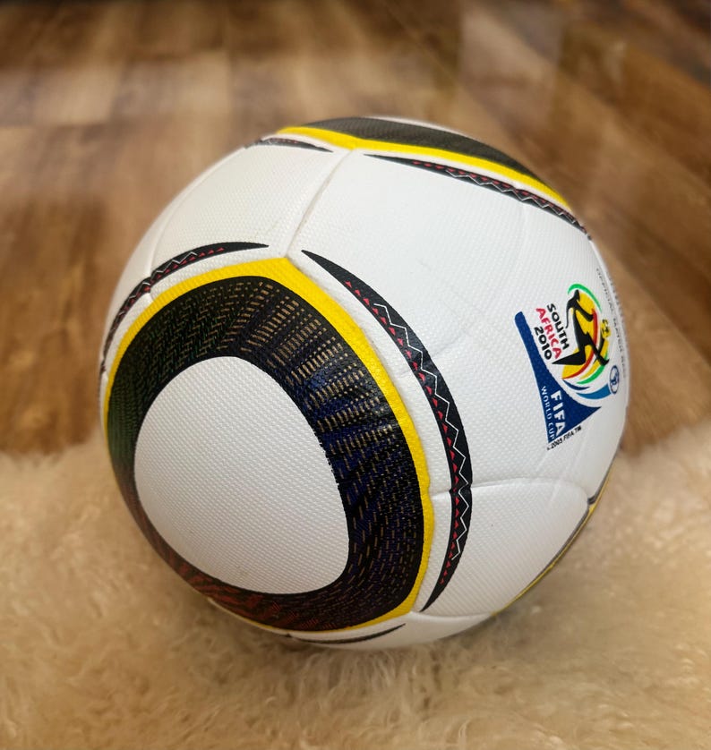K&ouml;nnte beinhalten: Ein wei&szlig;er Fu&szlig;ball mit schwarzen, gelben und roten Akzenten. Der Ball hat eine strukturierte Oberfl&auml;che und ein Design mit dem Text "SOUTH AFRICA 2010 FIFA WORLD CUP".