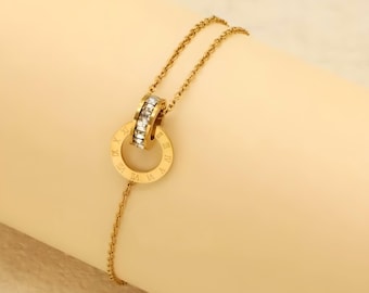 Könnte beinhalten: Goldfarbene Halskette mit einem kreisförmigen Anhänger mit römischen Ziffern und einem kleineren Ring, der mit klaren Kristallen besetzt ist. Die Halskette wird vor einem hellen Hintergrund präsentiert.