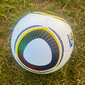 K&ouml;nnte beinhalten: Wei&szlig;er Fu&szlig;ball mit kreisf&ouml;rmigen Designelementen in Schwarz, Gelb, Gr&uuml;n und Rot. Der Ball hat ein FIFA-Logo und liegt auf gr&uuml;nem Gras. Er ist f&uuml;r die FIFA Fussball-Weltmeisterschaft 2010 konzipiert.