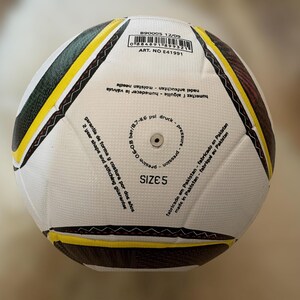 K&ouml;nnte beinhalten: Ein wei&szlig;-schwarzer Fu&szlig;ball mit gelben und braunen Akzenten. Der Ball hat die Gr&ouml;&szlig;e 5 und Text in mehreren Sprachen, darunter "2 Jahre Form- und Nahtgarantie".