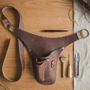 May include: A dark brown leather tool holster with an adjustable strap, metal D-rings, and rivets. The holster features a central pocket and a small side loop. Beside it lie a dark wooden stylus, a brass stitching chisel, an awl, and metal snips.