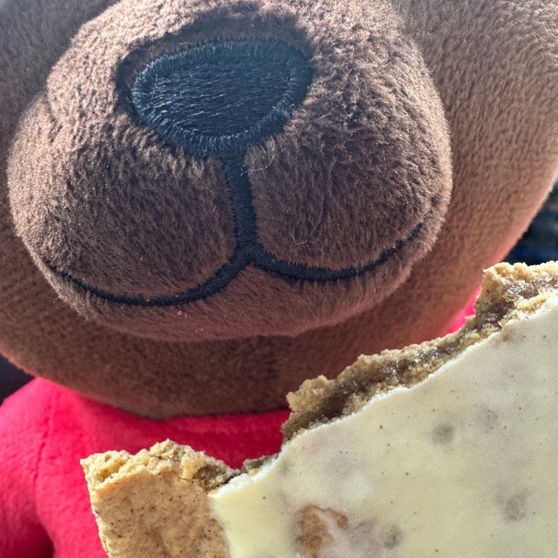 May include: A close-up of a brown teddy bear's face with a black stitched nose and mouth. The bear is wearing a red shirt. A partially eaten ice cream sandwich with a white filling is in the foreground.