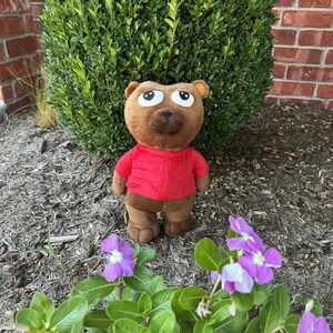 May include: A brown teddy bear wearing a red shirt and brown pants stands in front of a green bush. The bear has large, expressive eyes and a friendly expression. Purple flowers with white centers are in the foreground.