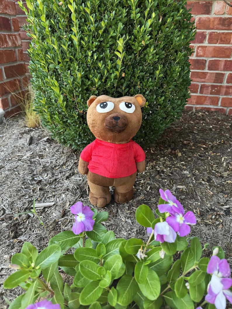 May include: A brown teddy bear wearing a red shirt and brown pants stands in front of a green bush. The bear has large, expressive eyes and a friendly expression. Purple flowers with white centers are in the foreground.