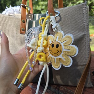 May include: A beige canvas tote bag with brown leather handles and a decorative keychain. The keychain features a yellow daisy with a smiling face, yellow cord, and clear beaded accents. The bag has a striped interior.