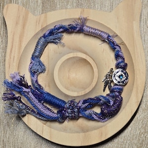 May include: A handcrafted, braided necklace in shades of blue and purple, accented with metallic threads. The necklace features a decorative silver charm with a blue gemstone and small angel wings. The necklace is displayed on a wooden, cat-shaped tray.