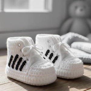 Könnte beinhalten: Ein Paar weiße Baby-Häkel-Sneakers mit schwarzen Streifen an den Seiten. Die High-Top-Schuhe haben weiße Schnürsenkel. Die Schuhe sind handgefertigt.