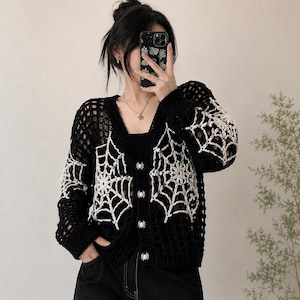May include: A black crochet cardigan featuring a V-neckline, long sleeves, and white spider web patterns on the front and sleeves. The garment is fastened with four silver-toned spider-shaped buttons down the centre front.