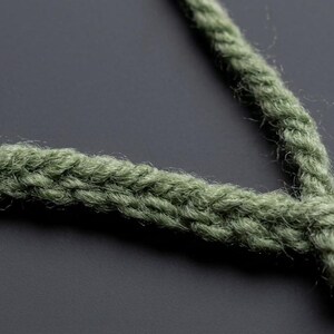 May include: A sage green crocheted cord or strap featuring a dense, braided-like stitch pattern. The yarn has a slightly fuzzy texture, and the cord is shaped into a V-formation, showcasing the consistent, interlocking loops of the handmade construction.