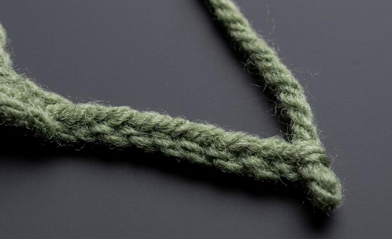 May include: A sage green crocheted cord or strap featuring a dense, braided-like stitch pattern. The yarn has a slightly fuzzy texture, and the cord is shaped into a V-formation, showcasing the consistent, interlocking loops of the handmade construction.