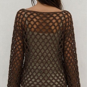 May include: A dark brown long-sleeve top featuring an open-knit crochet pattern with a diamond-shaped mesh design. The garment has a wide neckline and is layered over a light beige tank top, paired with light-colored pants.