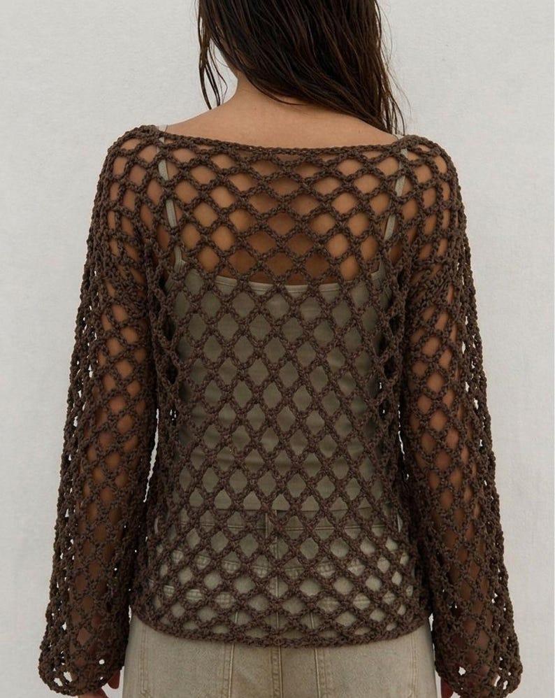 May include: A dark brown long-sleeve top featuring an open-knit crochet pattern with a diamond-shaped mesh design. The garment has a wide neckline and is layered over a light beige tank top, paired with light-colored pants.