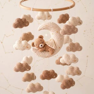 Può includere: Mobile per bambini fatto a mano con un orsetto all'uncinetto che dorme su una luna crescente, circondato da nuvole all'uncinetto nei toni del marrone e del crema. Il mobile è sospeso a un cerchio di legno.