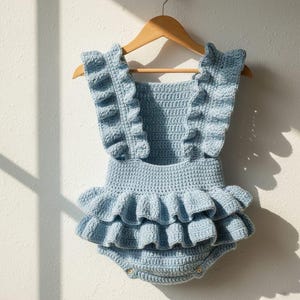 Könnte beinhalten: Hellblaue, handgehäkelte Baby-Spielanzug mit Rüschen an den Trägern und einem gestuften Rüschensaum. Das Kleidungsstück ist auf einem Holzbügel vor einer weißen Wand ausgestellt, was die filigrane Häkelarbeit und das Design hervorhebt.