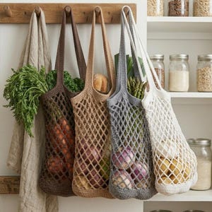 May include: Four crocheted net bags in shades of brown, tan, gray, and white, each filled with fresh produce. The bags hang from a wooden rack, showcasing carrots, bread, asparagus, and other vegetables. A linen towel hangs on the left.