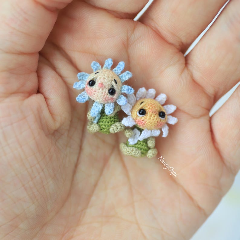 Puede incluir: Dos mu&ntilde;ecas de flores de ganchillo hechas a mano. Una mu&ntilde;eca tiene una flor azul claro con una cara melocot&oacute;n y hojas verdes. La otra mu&ntilde;eca tiene una flor blanca con una cara melocot&oacute;n y hojas verdes. Las mu&ntilde;ecas son peque&ntilde;as y detalladas.
