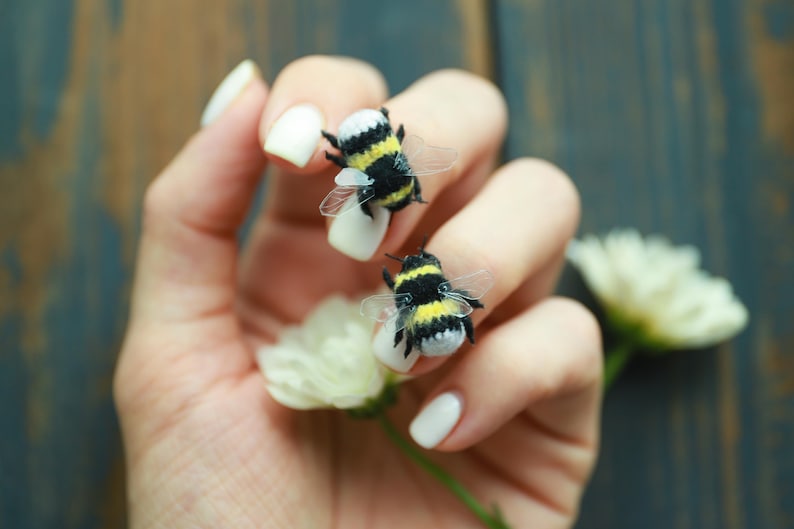May include: Two miniature bumblebee figurines with black and yellow stripes and transparent wings rest on white fingernails. A white flower and a blue-green wooden background complete the scene.