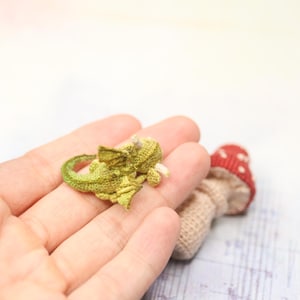 Tiny green micro crochet dragon resting on fingers, handmade amigurumi miniature with curled tail, small wings and horns, sleeping fantasy creature made with fine thread, scale shown by human hand.