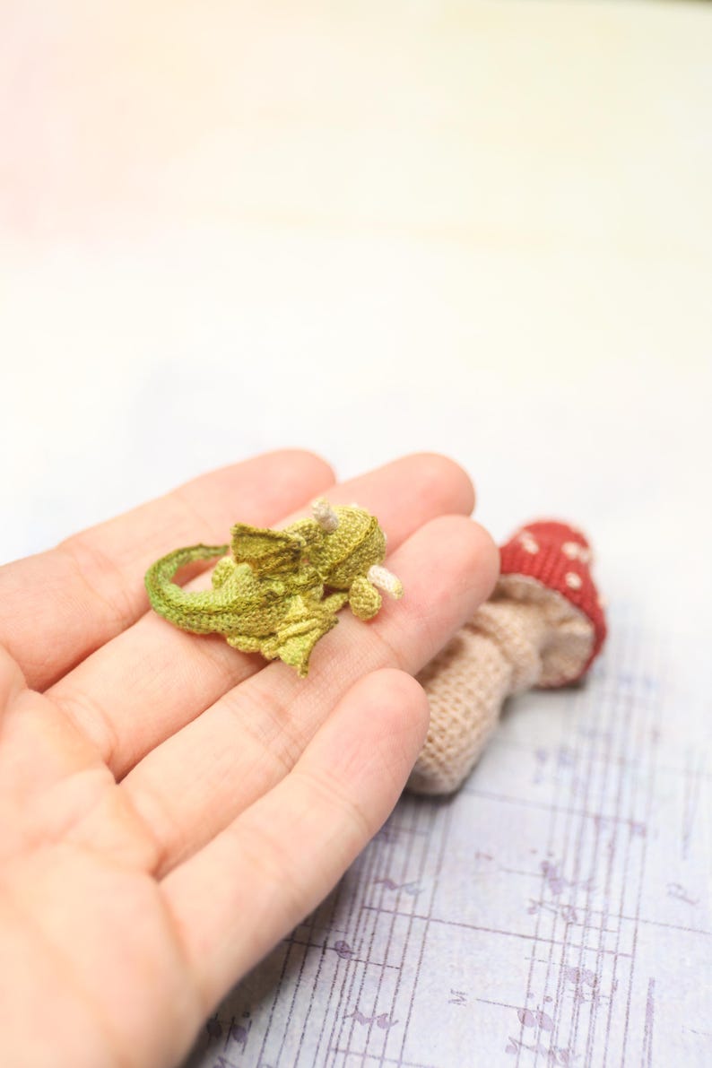 Tiny green micro crochet dragon resting on fingers, handmade amigurumi miniature with curled tail, small wings and horns, sleeping fantasy creature made with fine thread, scale shown by human hand.