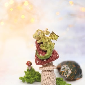 Miniature sleeping crochet dragon perched on crocheted red mushroom ring, tiny green amigurumi fantasy creature with folded wings and curled tail, detailed micro crochet toy shown on finger for scale.