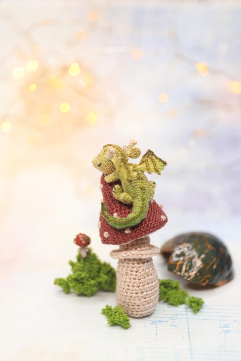 Miniature sleeping crochet dragon perched on crocheted red mushroom ring, tiny green amigurumi fantasy creature with folded wings and curled tail, detailed micro crochet toy shown on finger for scale.