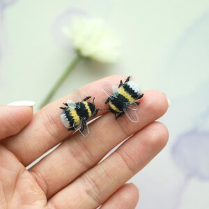 May include: Two handmade bee-shaped ornaments. The bees have black and yellow striped bodies, white and black heads, and transparent wings. They are resting on a person's hand, with a blurred flower in the background.