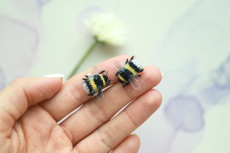May include: Two handmade bee-shaped ornaments. The bees have black and yellow striped bodies, white and black heads, and transparent wings. They are resting on a person's hand, with a blurred flower in the background.