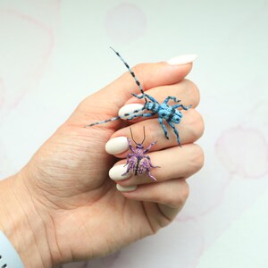 May include: Two miniature insect sculptures. One is blue with black markings, the other is purple with yellow accents. The insects are resting on a hand with white nail polish. A light blue watch with a gold buckle is visible.