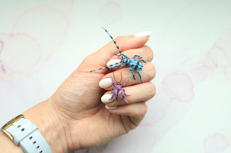 May include: Two miniature insect sculptures. One is blue with black markings, the other is purple with yellow accents. The insects are resting on a hand with white nail polish. A light blue watch with a gold buckle is visible.