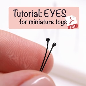 Peut inclure: Gros plan sur deux épingles à tête noire, probablement pour la fabrication de jouets miniatures. Les épingles sont tenues entre les doigts, sur un fond rose clair. Une bannière en haut indique "Tutoriel : YEUX pour jouets miniatures" avec une icône PDF.