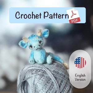 Puede incluir: Un pequeño dragón de ganchillo azul claro con cuernos beige y una expresión triste descansa sobre una bola de hilo gris. La imagen incluye una pancarta que dice "Crochet Pattern PDF" y una insignia de "English Version".