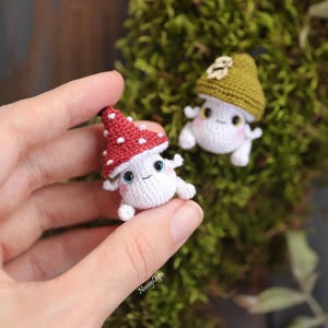 Pu&ograve; includere: Primo piano di due piccole figure di funghi all'uncinetto. Una &egrave; tenuta in mano, con un cappello rosso a pois bianchi. L'altra ha un cappello verde. Entrambi hanno facce sorridenti e piccoli arti. Lo sfondo &egrave; muschio verde.