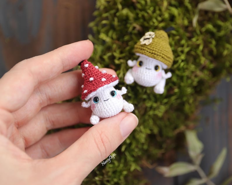 Pu&ograve; includere: Primo piano di due piccole figure di funghi all'uncinetto. Una &egrave; tenuta in mano, con un cappello rosso a pois bianchi. L'altra ha un cappello verde. Entrambi hanno facce sorridenti e piccoli arti. Lo sfondo &egrave; muschio verde.