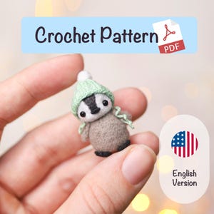 May include: A miniature crochet badger, wearing a green hat and scarf, held in a hand. The badger is grey and white with black features. The image includes the text "Crochet Pattern" and "English Version".