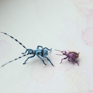 May include: Two handmade crochet insect figures. One is blue with black markings and long antennae. The other is a smaller, purple insect. They are set against a white background with pink watercolour splotches.