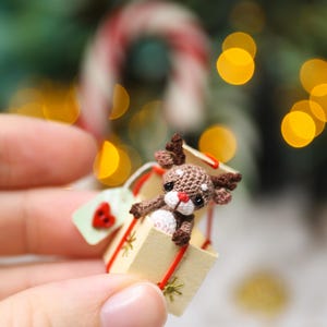May include: A miniature crocheted reindeer peeks out of a small gift box. The reindeer is brown with a white muzzle and red nose. The box is beige with a red ribbon and a small tag with a red heart. Festive holiday decor.