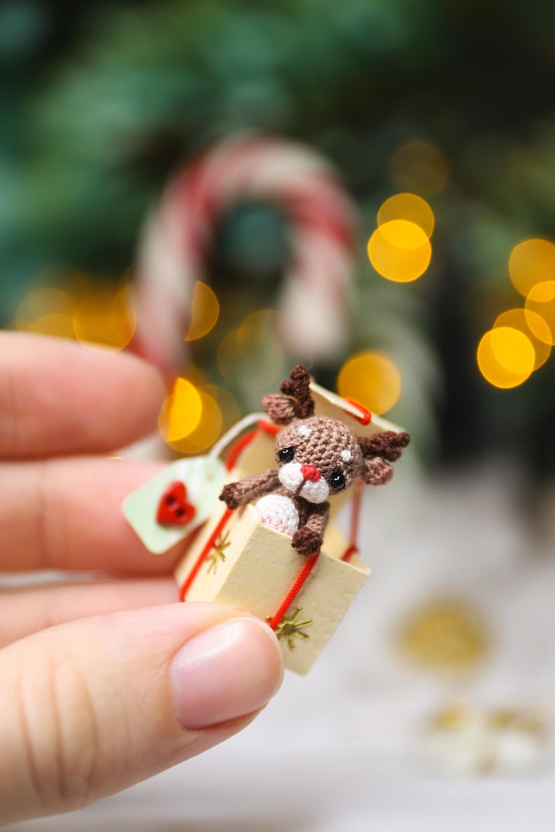 May include: A miniature crocheted reindeer peeks out of a small gift box. The reindeer is brown with a white muzzle and red nose. The box is beige with a red ribbon and a small tag with a red heart. Festive holiday decor.