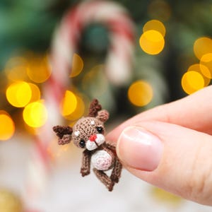 May include: A miniature crocheted reindeer with brown and white details, including antlers, a red nose, and a white belly. The tiny Christmas ornament is held in a hand, with a blurred background of festive lights and a candy cane.