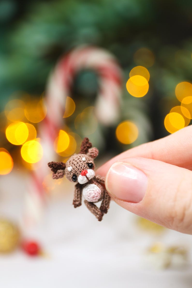 May include: A miniature crocheted reindeer with brown and white details, including antlers, a red nose, and a white belly. The tiny Christmas ornament is held in a hand, with a blurred background of festive lights and a candy cane.
