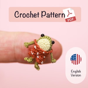 May include: A miniature crochet frog wearing red overalls with white polka dots, featuring black bead eyes and a cream-colored belly. The image includes text that reads Crochet Pattern PDF and English Version with a US flag icon.
