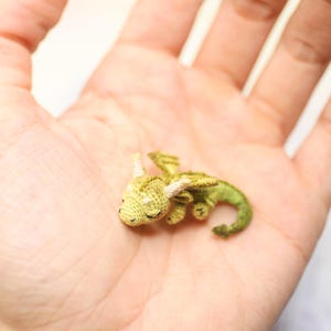 Tiny green micro crochet dragon resting on fingers, handmade amigurumi miniature with curled tail, small wings and horns, sleeping fantasy creature made with fine thread, scale shown by human hand.