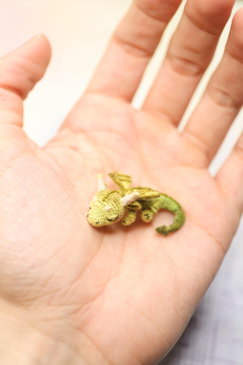Tiny green micro crochet dragon resting on fingers, handmade amigurumi miniature with curled tail, small wings and horns, sleeping fantasy creature made with fine thread, scale shown by human hand.