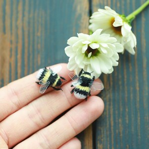 May include: Two miniature bumblebees with black and yellow stripes and transparent wings rest on a person's fingers. White flowers with yellow centers and green stems are in the background. The bees are a craft item.