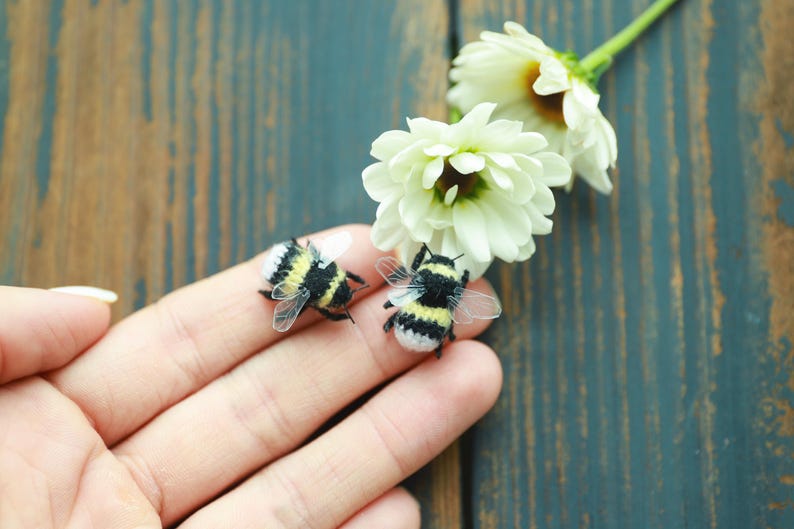 May include: Two miniature bumblebees with black and yellow stripes and transparent wings rest on a person's fingers. White flowers with yellow centers and green stems are in the background. The bees are a craft item.