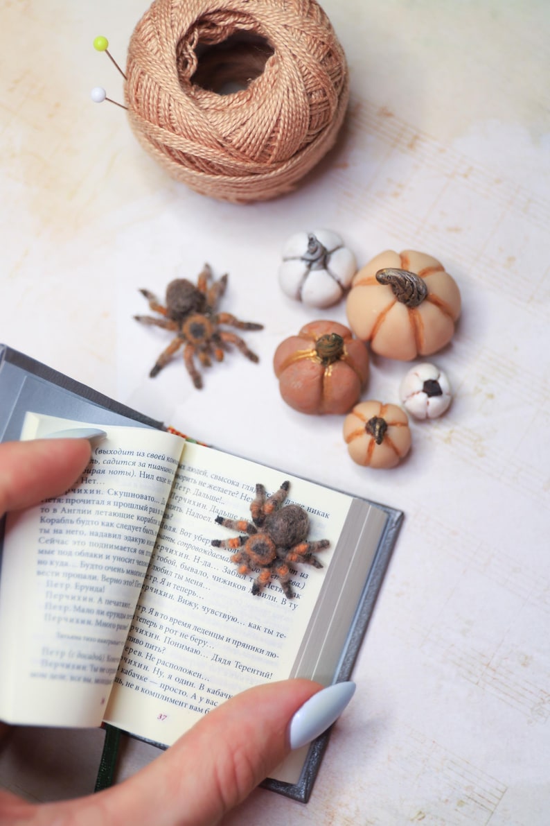 May include: A collection of miniature handmade pumpkins in various sizes and colors, including orange, white, and brown. A small, detailed spider figurine rests on an open book. A ball of tan yarn with pins is in the background.