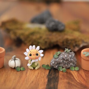 Puede incluir: Una escena de jard&iacute;n en miniatura caprichosa con un personaje de margarita de ganchillo, peque&ntilde;as macetas de terracota con plantas, una calabaza blanca y una roca con vegetaci&oacute;n. La escena est&aacute; sobre una superficie de madera.